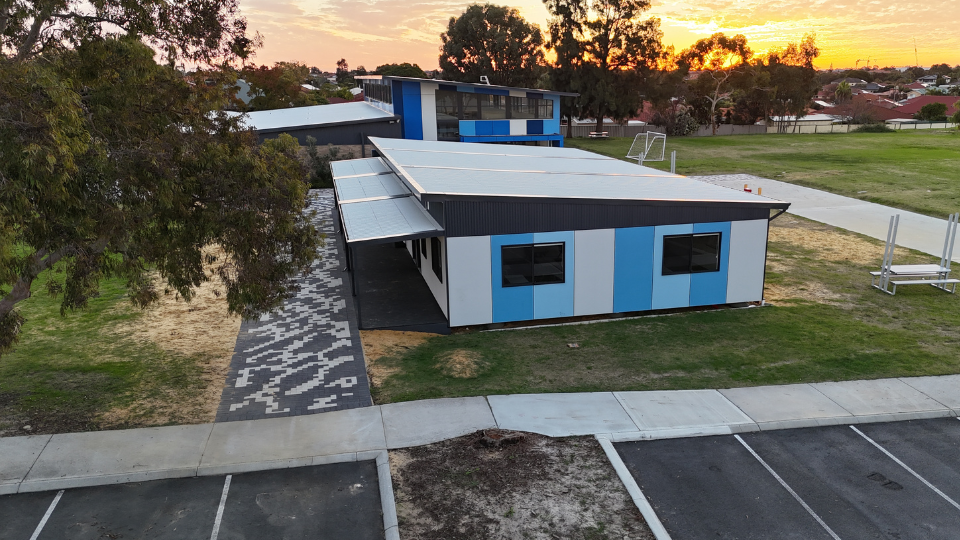 A sleek modular building with a modern white and blue panelled exterior, set within a landscaped school campus in Western Australia at sunset, featuring a patterned walkway, green open space, and parking nearby.