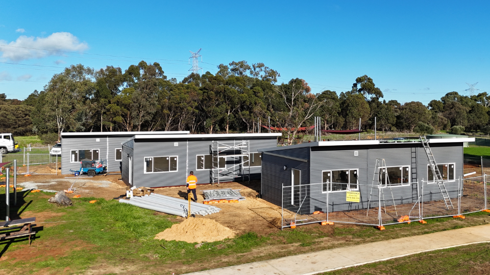 Three modular buildings nearing completion on a construction site in Western Australia, featuring grey cladding, large windows, and scaffolding, with construction workers and materials in the foreground and a backdrop of trees under a clear blue sky