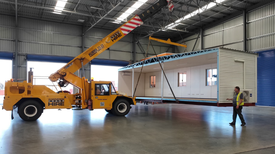 A prefabricated modular building being carefully lifted by a yellow mobile crane inside a spacious industrial warehouse, demonstrating off-site construction efficiency with a worker overseeing the process in Western Australia.